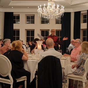 a man in a red check shirt stands with arms outstretched, adressing diners a round table at a formal dinner occasion. there is a crytal chandelier on the ceiling above him